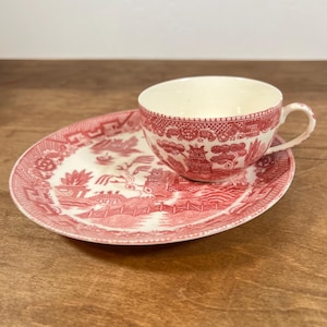May include: A cream-colored ceramic teacup and matching saucer featuring a detailed red transferware pattern of a landscape with pagodas, trees, and a bridge. The teacup has a curved handle and the saucer has a wide, decorative border.