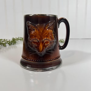 May include: A brown ceramic mug with a detailed fox head design. The mug has a silver rim and base, and a dark brown handle. The fox's fur is a mix of brown and orange tones, with detailed facial features.