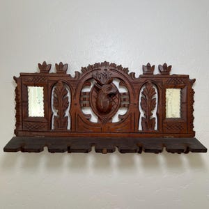 May include: A dark brown, carved wooden wall shelf with a deer head centerpiece. The shelf features two small mirrors and decorative leaf and crown accents. The shelf has a lower section with several small holes.