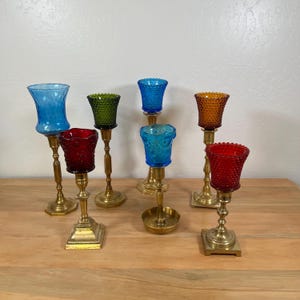 May include: A collection of six candle holders featuring brass stands and colorful glass votive cups. The glass cups include textured hobnail patterns in red, blue, green, and orange. Each holder has a distinct base shape, ranging from square to round, supporting the various glass inserts.