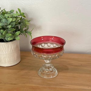 May include: A clear glass footed bowl with a ruby red rim. The bowl has a decorative pattern around the base and rim. The bowl is sitting on a wooden surface, with a potted plant in the background.