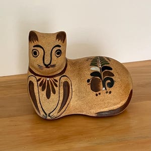 May include: A tan ceramic cat figurine with dark brown and black painted details. The cat has large eyes, a detailed collar, and decorative patterns on its body. The figurine is resting on a wooden surface.