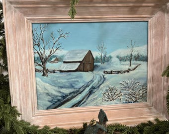 May include: A framed winter landscape painting featuring a snow-covered barn and a winding road. The artwork is set against a light blue sky, with leafless trees and a wooden fence. The frame is a light, neutral color.