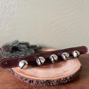 May include: A brown leather strap with six silver bells attached. The bells are evenly spaced along the strap. The strap is resting on a wooden slice, with pine needles and a pine cone in the background.