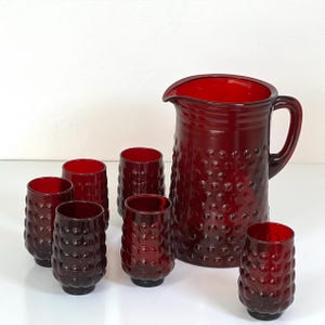 May include: A set of deep red glass drinkware consisting of one large pitcher and five matching tumblers. Each piece features a distinct hobnail texture with raised circular bumps covering the exterior surface, creating a tactile pattern.