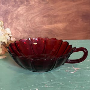 May include: A ruby red glass teacup with a handle. The cup has a scalloped edge and a flower-like shape. The cup is sitting on a light green surface.