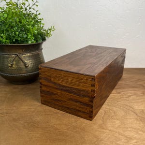 May include: A rectangular wooden box with a dark brown finish and visible wood grain. The box features interlocking finger joints at the corners, creating a secure construction for the lid and base. The surface is smooth and polished.