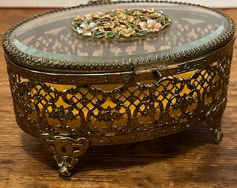May include: Oval-shaped vintage jewelry box with a glass top and ornate gold-colored metal filigree. The lid features a floral design with pink, blue, and white accents. The interior is lined with yellow fabric, and the box rests on decorative feet.