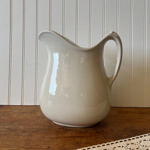 May include: A cream-colored ceramic pitcher with a curved handle and a pouring spout. The pitcher has a rounded body and a smooth, glossy finish. The background is a white paneled wall and a wooden surface.