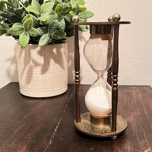 May include: An hourglass with a clear glass bulb filled with white sand, set in a brass frame. The hourglass has a decorative base and top, and is displayed on a dark wooden surface. A potted plant is in the background.