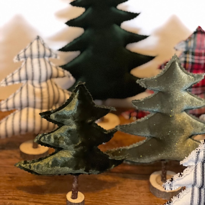 Pu&ograve; includere: Un set di cinque alberi di Natale in miniatura, ognuno con un tessuto e un colore diversi. Gli alberi sono fatti di feltro e hanno una base in legno rustica. Gli alberi sono disposti in fila su una superficie in legno.