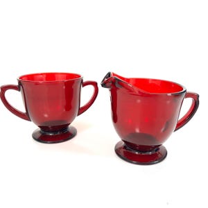 May include: A ruby red glass creamer and sugar bowl set. The creamer has a spout and handle, while the sugar bowl has two handles. Both pieces have a rounded base and are made of transparent glass.