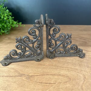 May include: Two dark brown cast iron shelf brackets featuring an intricate scrollwork design with openwork patterns. Each bracket has a triangular shape with mounting holes at the base and along the vertical edge for secure wall installation.