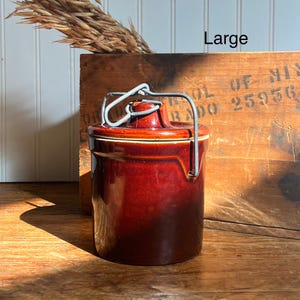 May include: A vintage, dark brown ceramic crock with a lid and metal handle. The crock has a glossy finish and is labeled "Large". The background includes a wooden crate and dried plants.
