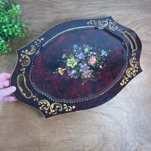 May include: An oval serving tray with a deep burgundy and black marbled finish, featuring gold scrollwork along the edges and a central floral bouquet painting with yellow, purple, pink, and blue flowers. It includes two integrated handles.