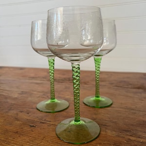May include: Three clear wine glasses with green stems and bases. The stems have a twisted design. The glasses are on a wooden surface with a white background.
