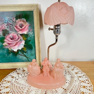 May include: A vintage pink table lamp with a decorative base featuring three figures. The lamp has a curved brass stem and a pink, textured lampshade. A framed painting of pink roses is in the background.
