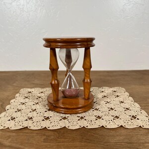 May include: A wooden hourglass with a polished brown finish, featuring a clear glass bulb containing fine pink sand. The frame consists of a circular top and base connected by four turned spindles, creating a classic timekeeping instrument.