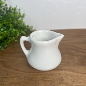 May include: A small, white ceramic creamer pitcher with a rounded body, a curved handle on the left side, and a small pouring spout on the right side. The pitcher has a smooth, glossy finish and a simple, functional design for serving cream or milk.