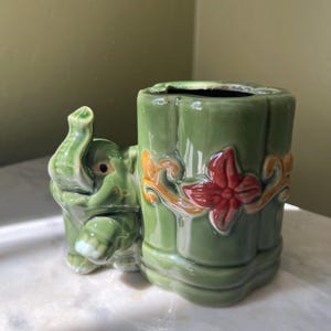 May include: A green ceramic elephant planter with a raised trunk, attached to a cylindrical container. The container has a red flower and orange accents. The planter is on a white surface.