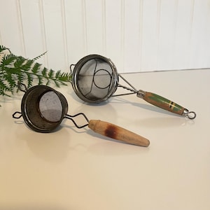 May include: Two vintage metal tea strainers with wooden handles. One strainer has a green and gold painted handle, the other has a natural wood handle. Both strainers have a fine mesh screen and are designed for steeping tea leaves.
