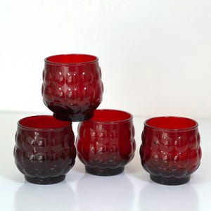May include: A set of four deep red glass tumblers featuring a textured, raised circular bubble pattern on the exterior. Each tumbler has a rounded, bulbous shape with a smooth rim and a flat base, designed for holding beverages or as decorative items.