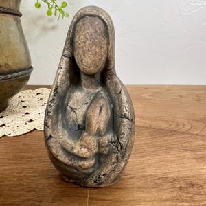 May include: A textured, bronze-toned sculpture depicting a stylized figure with a hooded head and draped clothing. The surface features a weathered, cracked finish with dark brown and metallic gold highlights, creating an abstract, organic form.