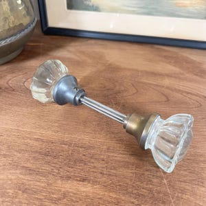 May include: A door knob set featuring two clear, fluted glass knobs connected by a central metal spindle. Each knob is attached to a circular metal base, with one base appearing silver-toned and the other showing a darker, aged brass finish.