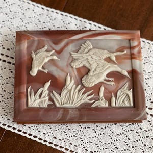 May include: A rectangular box with a marbled reddish-brown and white finish. The lid features a raised, cream-colored relief carving of three flying ducks soaring above a cluster of cattail plants and reeds, set against a smooth, swirling background.