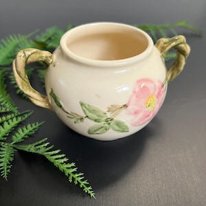 May include: Cream-colored ceramic sugar bowl with a pink floral design and green leaves. The bowl has two twisted handles and a round shape. The floral design is on the side of the bowl.