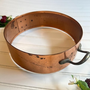 May include: A copper-colored, circular cooking mold with a handle. The mold has a slightly aged appearance, with some darker spots on the surface. The handle is a darker metal. The mold is designed for baking or cooking.