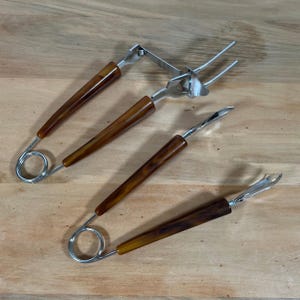 May include: Set of four vintage serving utensils with brown, marbled handles and stainless steel components. Includes two forks, a spoon, and a serving tool with a spring handle. The utensils are arranged on a light brown wooden surface.