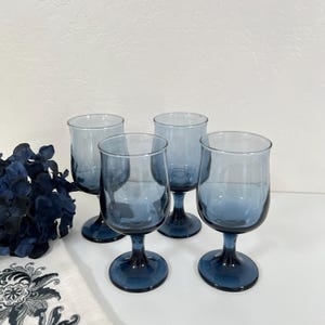 May include: Four blue-tinted wine glasses with a rounded bowl and a short stem. The glasses are arranged on a white surface, with a dark blue floral arrangement and a patterned cloth in the background.