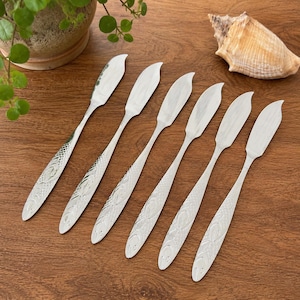 May include: A set of six silver-toned butter knives arranged in a row. Each knife features a pointed blade and a handle decorated with an embossed fish scale pattern. The metal surface has a polished finish, reflecting light across the textured handles.
