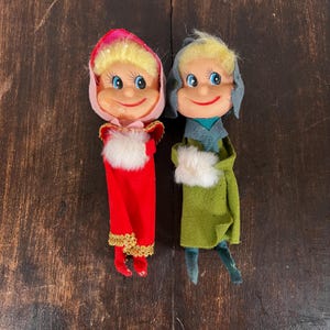 May include: Two vintage Christmas elf dolls. One doll is wearing a red outfit with gold trim and a pink hat. The other doll is wearing a green outfit with a blue scarf and hat. Both dolls have blonde hair and smiling faces.