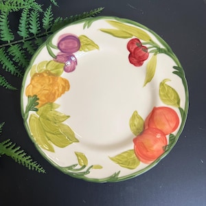 May include: A white ceramic plate with a green rim and a colorful fruit and leaf design. The plate features illustrations of purple grapes, red tomatoes, and orange peaches, with green leaves and vines.