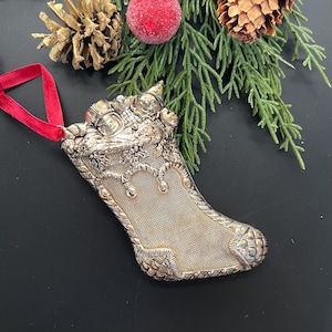 May include: A silver Christmas stocking ornament with intricate detailing, including a decorative top with figures and holly. The ornament is suspended by a red ribbon and is approximately 4 inches long.