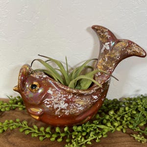 May include: A ceramic fish-shaped planter with a glossy, iridescent copper-brown glaze and textured white and gold accents. The fish has a single dark eye and an open mouth, containing a green air plant with thin, pointed, arching leaves.