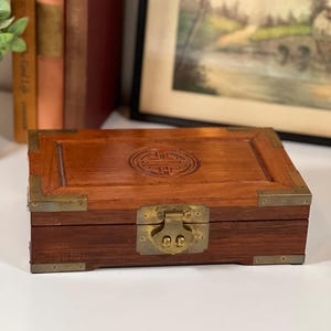 May include: A rectangular wooden box with a hinged lid, brass hardware, and a decorative emblem on the top. The box is a warm brown color and appears to be vintage or antique. The box is closed with a brass clasp.