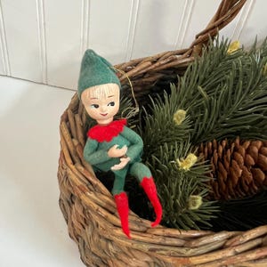 May include: A vintage Christmas elf ornament with a green hat and suit, red collar and boots, and a painted face. The elf is seated in a woven basket with pine branches and a pine cone.
