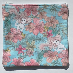 May include: A blue zippered pouch with a white zipper. The pouch is decorated with a floral pattern and images of mermaids. The mermaids are drawn in a cartoon style and have long flowing hair.