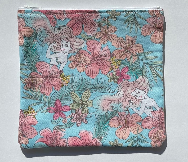 May include: A blue zippered pouch with a white zipper. The pouch is decorated with a floral pattern and images of mermaids. The mermaids are drawn in a cartoon style and have long flowing hair.