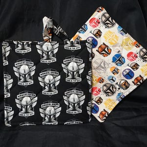 May include: Two quilted pot holders. One is black with a repeating pattern of a Mandalorian helmet and the text "THIS IS THE WAY". The other is white with a repeating pattern of colorful Mandalorian helmets.