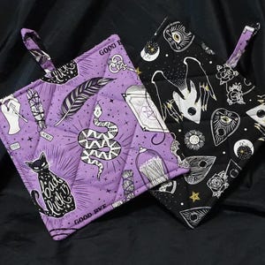 May include: Two quilted pot holders with hanging loops. One is lavender with a snake, cat, and feather design. The other is black with a celestial and Ouija board design. Both have the words "Good Bye" and "Bad Luck" printed on them.