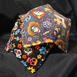 May include: Two fabric bowl cozies with a black background and colorful patterns featuring characters from the movie Coco. The cozies have a purple and black pattern with a skeleton and a floral pattern with a skull.