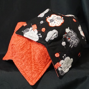 Puede incluir: Una almohada de tela negra y roja con un diseño de ovejas blancas y grises. Las ovejas sostienen ovillos de lana y agujas de tejer. La almohada es reversible con un patrón de rayas rojas y blancas en el otro lado.