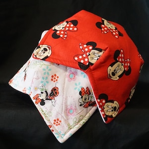May include: A set of two red and white pot holders featuring a Minnie Mouse design. The red pot holder has a solid red background with white polka dots and Minnie Mouse faces. The white pot holder has a white background with a floral pattern and Minnie Mouse figures.