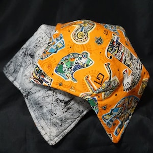 Puede incluir: Dos posavasos de tela, uno con un patrón geométrico blanco y gris y el otro con un fondo naranja brillante y un estampado de animales abstracto y colorido.