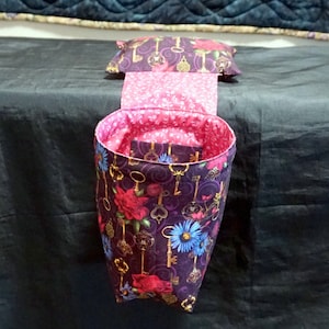 May include: A purple fabric hanging organizer with a pink lining and a keyhole pattern. The organizer has a pocket on the front and is filled with gold keys and roses.