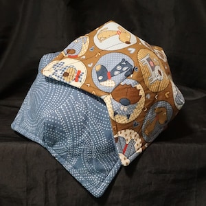 May include: A reversible bowl cozy featuring a blue and white dotted pattern on one side and a brown fabric with dog illustrations on the other. The cozy is square-shaped and designed to hold a bowl of food.